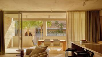 A modern living room with a sofa, a dining table with chairs, and a view through a glass door to an outdoor area with plants and a brick wall.