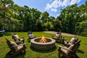 A circular fire pit surrounded by four Adirondack chairs with cushions, set in a grassy outdoor area with trees in the background.