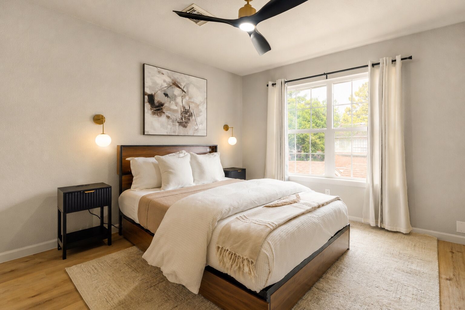 A bedroom with a bed, bedside tables, a ceiling fan, a window with curtains, and a wall-mounted artwork.