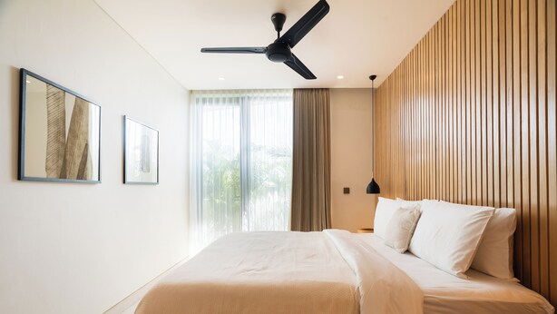 A modern bedroom with a bed, ceiling fan, large window, and wooden panel wall.
