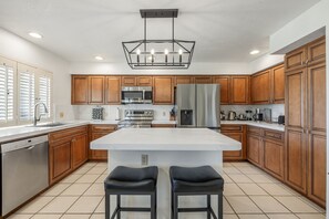 A modern kitchen with wooden cabinets, stainless steel appliances, a central island with stools, and a pendant light. - 4BR Desert Retreat on the Golf Course Desert Falls (Palm Desert)