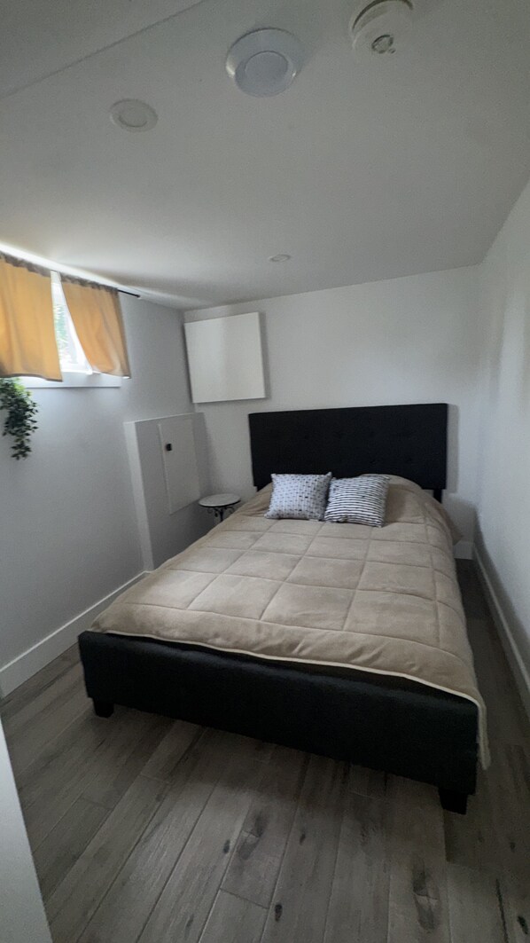 A bedroom with a bed, a headboard, a bedside table, a lamp, and a window with curtains.