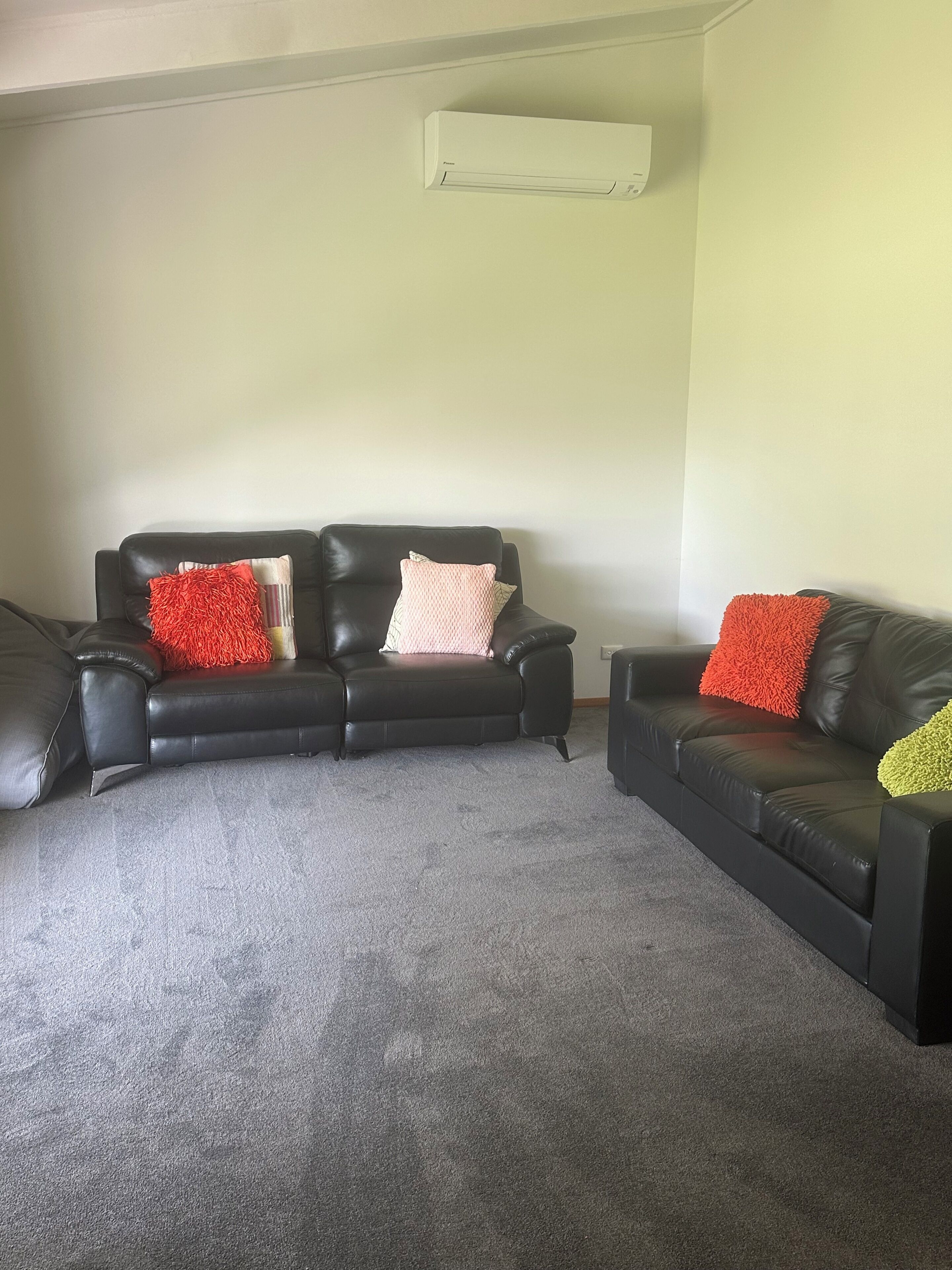 A living room with a black leather sofa, a grey carpet, and a white air conditioning unit on the wall.