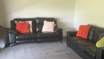 A living room with a black leather sofa, a grey carpet, and a white air conditioning unit on the wall.
