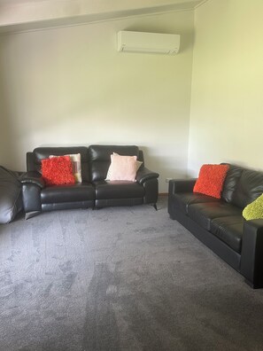 A living room with a black leather sofa, a grey carpet, and a white air conditioning unit on the wall.