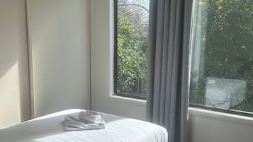 A neatly made bed with a white comforter and folded towels, a window with curtains, and a view of greenery outside.