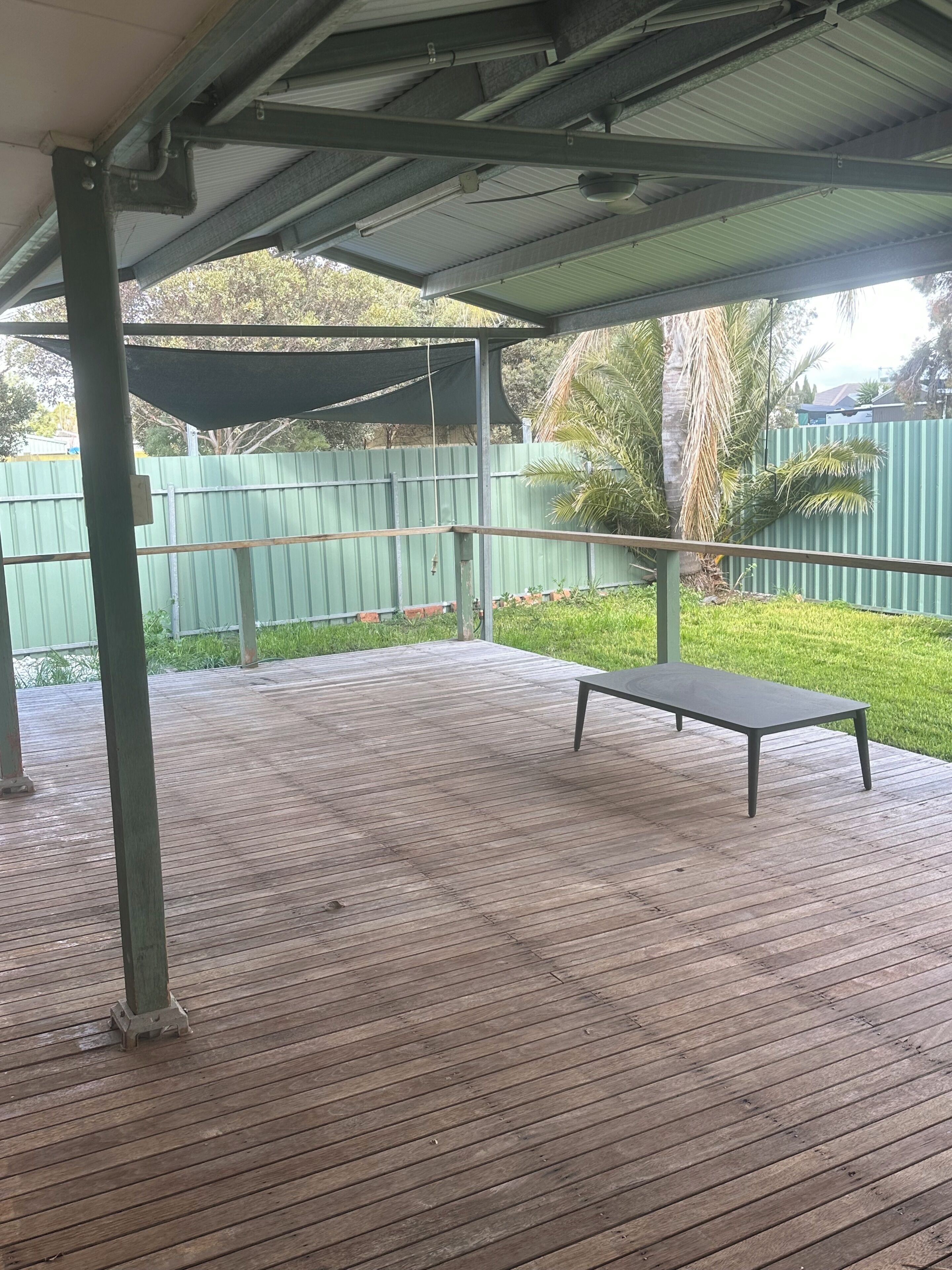A covered outdoor area with a wooden deck, a metal table, and a green fence in the background.