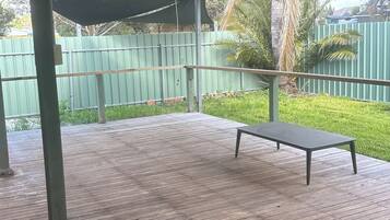 A covered outdoor area with a wooden deck, a metal table, and a green fence in the background.