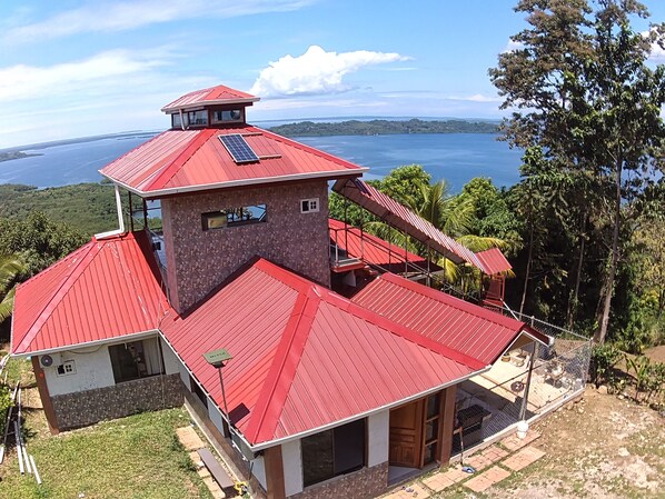 A two-story building with a red roof, a small tower with a balcony, and a view of a lake.
