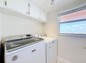 A laundry room with a front-loading washer and dryer, white cabinets above, and a window with a view. - Ocean View Beach Condo w/ Heated Pool in Cocoa Beach (Cocoa Beach)