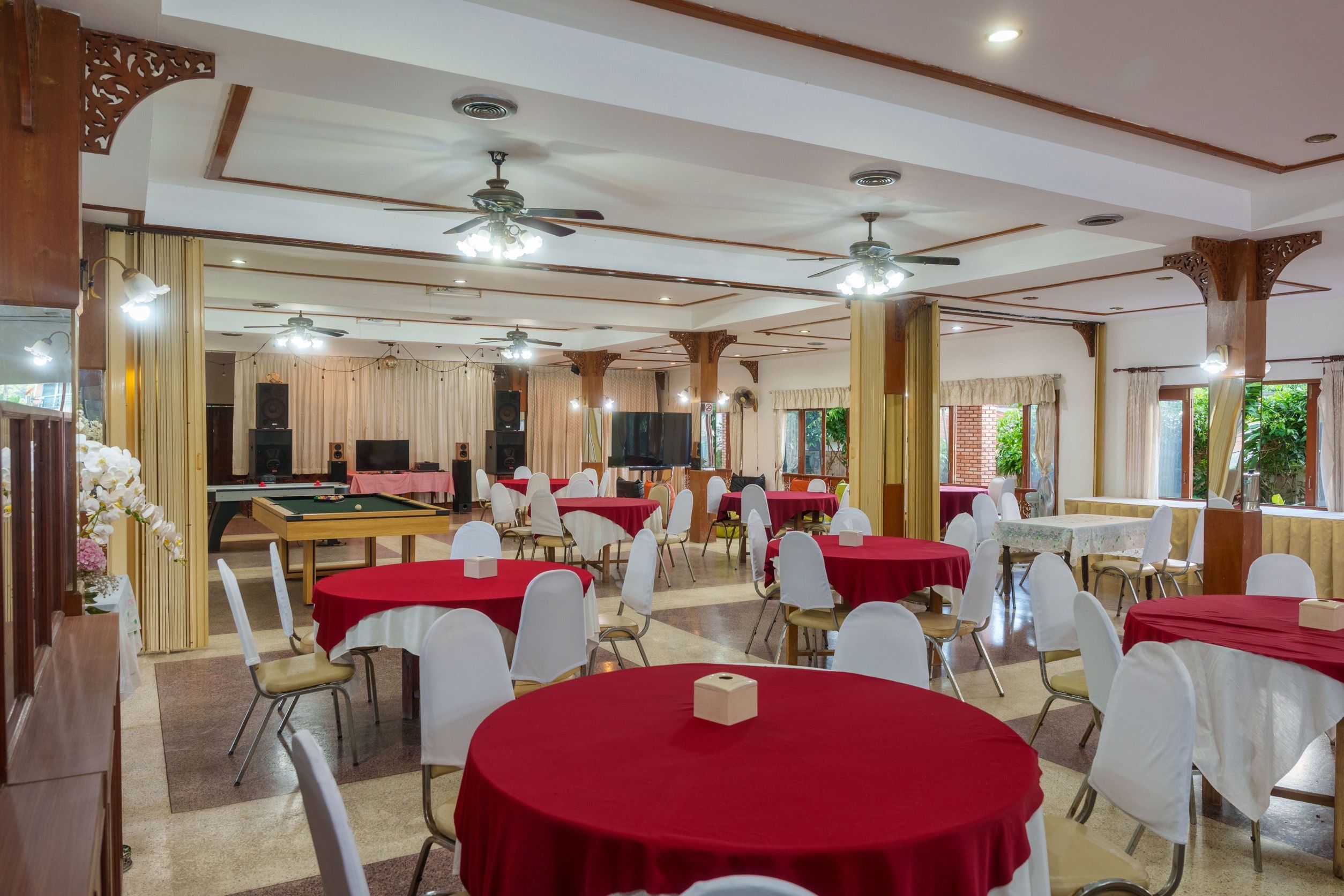 Een feestzaal met meerdere ronde tafels, bedekt met rode tafelkleden, witte stoelen en plafondventilatoren.