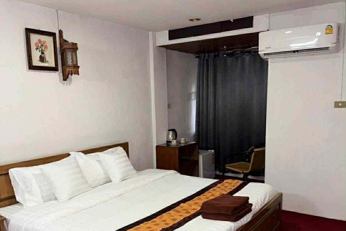 Habitación de hotel con una cama individual, una mesita de noche, una silla, un escritorio, un marco de fotos, aire acondicionado instalado en la pared y una cortina.
