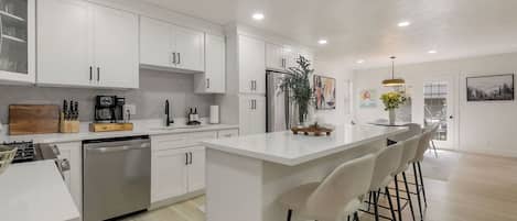 A modern kitchen with white cabinetry, a central island with bar stools, and a dining area with a view.