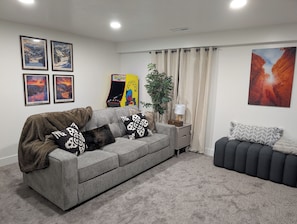 Ein Wohnzimmer mit grauem Sofa, gemusterten Kissen, einem kleinen Beistelltisch mit Lampe und einer Pflanze.
