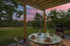 An outdoor patio with a round table set for dining, surrounded by chairs, and a barbecue grill in the background.