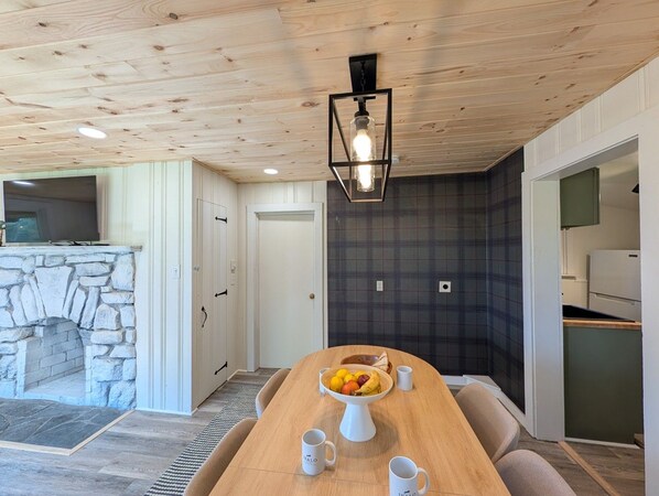 Un comedor con chimenea de piedra, una mesa de madera con frutero y una lámpara colgante moderna.