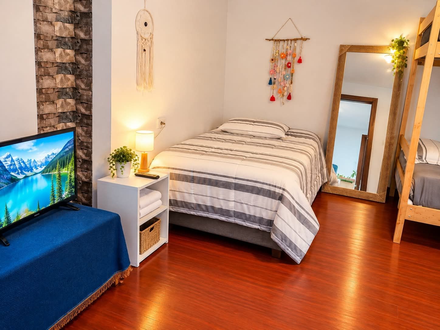 Ein Schlafzimmer mit einem Bett, einem Fernseher, einem Nachttisch mit Lampe, einem Wandbehang im Traumfänger-Stil und einem Spiegel.