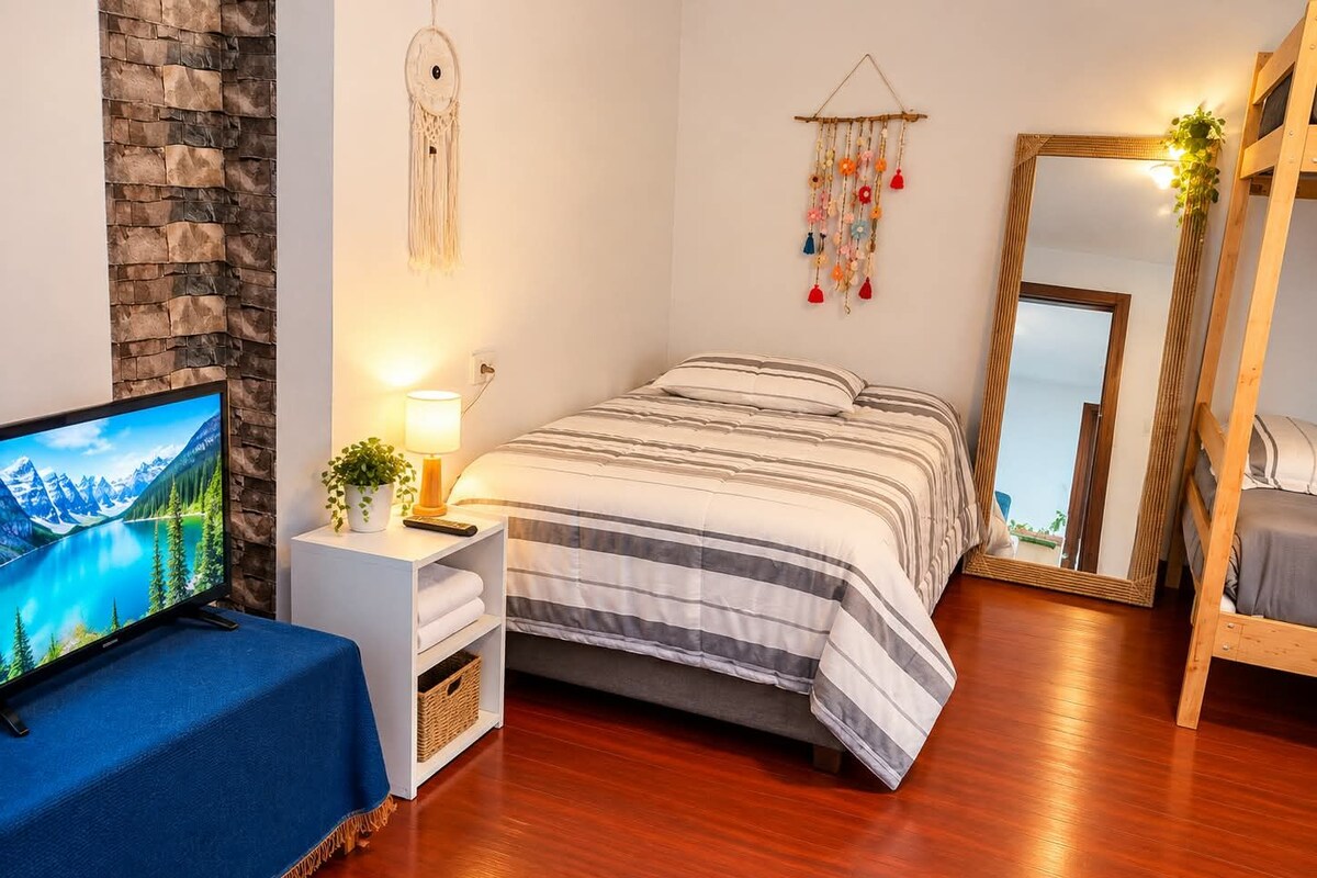A bedroom with a bed, a TV, a bedside table with a lamp, a wall-mounted dreamcatcher, and a mirror.