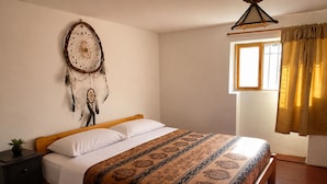 Un dormitorio con cama de madera, ropa de cama blanca, un atrapasueños en la pared, una planta pequeña en una mesita de noche y una ventana con cortinas.