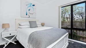 Un dormitorio con cama, mesita de noche, lámpara, ventana con vista a árboles y un cuadro abstracto colgado en la pared.