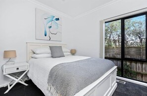 Ein Schlafzimmer mit einem Bett, einem Nachttisch, einer Lampe, einem Fenster mit Blick auf Bäume und einem an der Wand hängenden abstrakten Gemälde.
