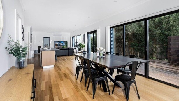 Un comedor moderno con una mesa larga y sillas, una cocina al fondo, y grandes ventanales que ofrecen vistas al exterior.