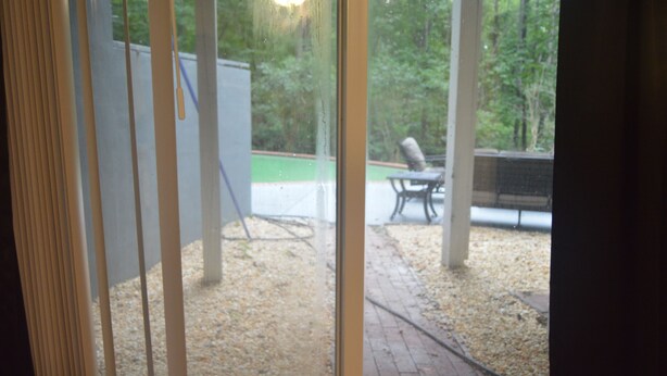 Une vue à travers une fenêtre donnant sur un patio, avec une table et des chaises, et un espace vert en arrière-plan.