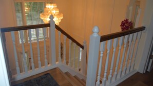 Un escalier avec des balustrades blanches et une main courante en bois, menant à une pièce dotée d’un lustre et d’une fenêtre avec des stores.