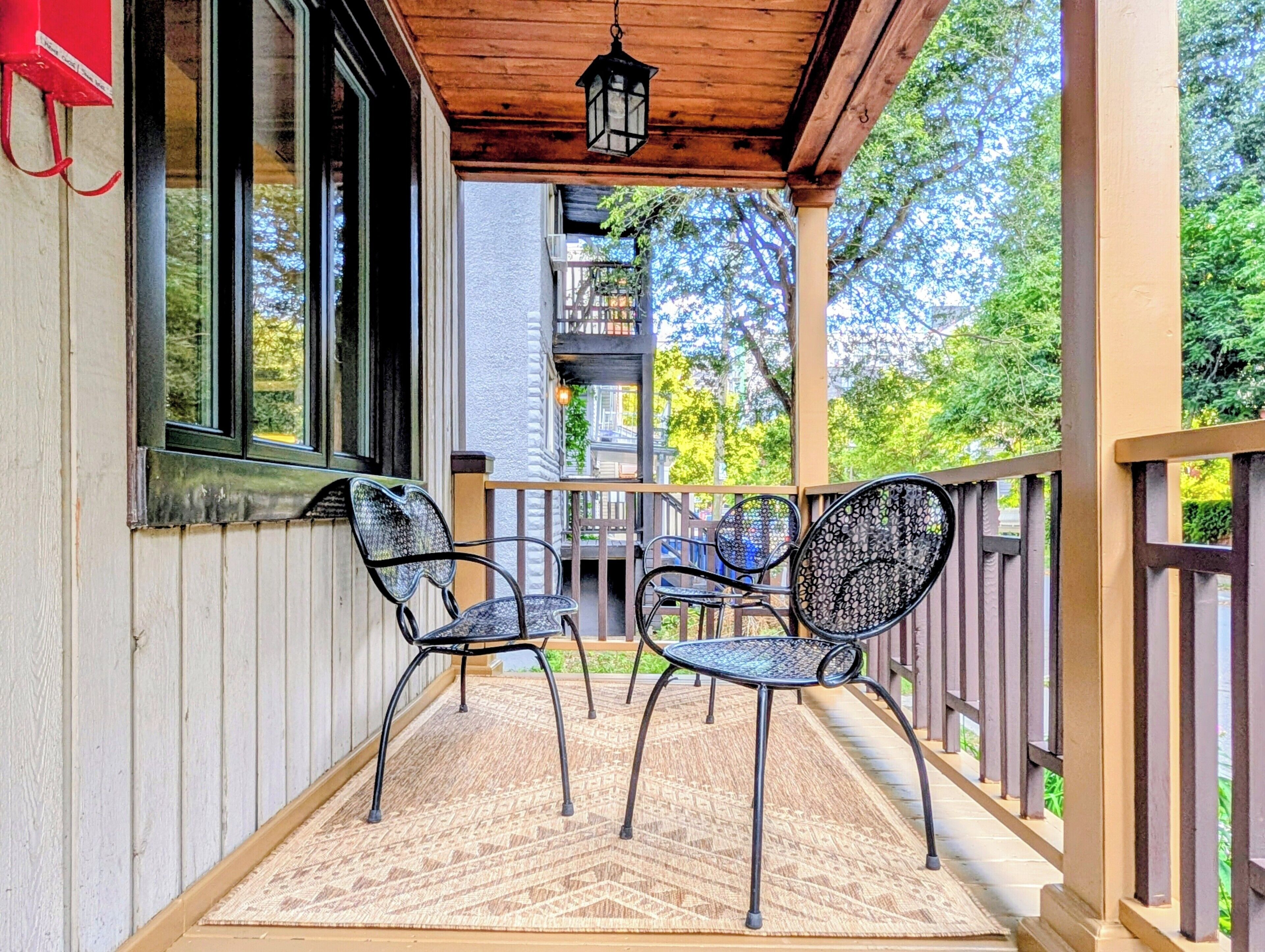 Une véranda avec deux chaises et une table, une balustrade en bois et une vue sur un bâtiment en arrière-plan.