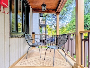 A covered porch with two chairs and a table, a wooden railing, and a view of a building in the background.