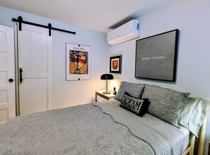 Une chambre à coucher comprenant un lit, une table de chevet, une lampe, un climatiseur fixé au mur et des œuvres d’art encadrées.