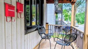 A wooden porch with two metal chairs and a rug, a wooden railing, and a view of a street and trees. - Luxury Duplex in a Prime Location (Gatineau)