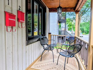 A wooden porch with two metal chairs and a rug, a wooden railing, and a view of a street and trees. - Luxury Duplex in a Prime Location (Gatineau)