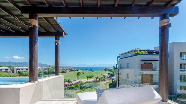 Un balcon avec vue sur un terrain de golf et l’océan, des fauteuils de jardin en bois et une structure en forme de pergola.