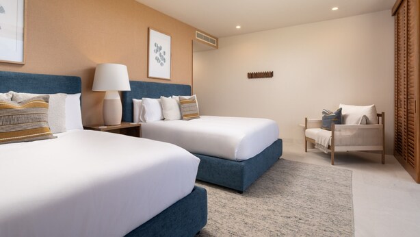 Een hotelkamer met twee bedden, een nachtkastje met lamp, een stoel en kunstwerken aan de muur.