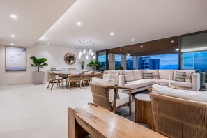 A modern living room with a large sectional sofa, dining area, and a view of the cityscape through large windows. - Amor y Surf 5BD - Surf Punta Mita (Punta de Mita)