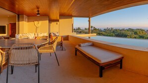 Amplia habitación con techo alto, zona de comedor con mesa y sillas, espacio de estar con banco, y un gran ventanal con vistas a palmeras y montañas.