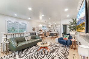 A modern living room with a green sofa, a zebra-patterned rug, and a dining area with a kitchen in the background. - Honey's Full House-Both Homes, Hot Tub, Sleeps 12 (Pocatello)