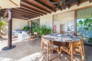 A modern outdoor dining area with a round table and wooden chairs, surrounded by potted plants and a covered seating area with a sofa and chairs.