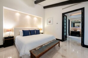 A modern bedroom with a large bed, bedside lamps, a bench, and a view into a bathroom. - Condo Jazmin 4BD - Hacienda De Mita Punta Mita (Punta de Mita)
