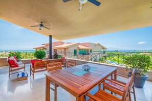 Ein Balkon mit Holztisch und -stühlen, einem Deckenventilator und Blick auf das Meer und Gebäude.