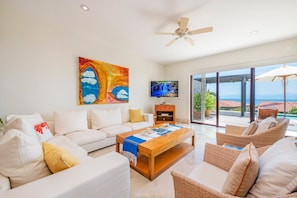 A spacious living room with a white sofa, a wooden coffee table, and wicker chairs. A ceiling fan is visible, and there is a large window offering a view of the ocean. - Villa Perla 3BD - Porta Fortuna Punta Mita (Punta de Mita)