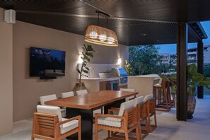 Ein Essbereich im Freien mit einem Holztisch und Stühlen, einer hängenden Lampe, einem an der Wand befestigten Fernseher und einem Grill.