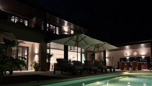 En pool med solstolar och parasoller, en matplats med bord och stolar samt en modern byggnad med stora fönster.