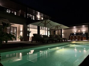 Ein Schwimmbecken mit Liegestühlen und Sonnenschirmen, ein Essbereich mit Tischen und Stühlen sowie ein modernes Gebäude mit großen Fenstern.