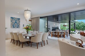 Ein moderner Essbereich mit einer Chandelüster, einem großen Esstisch für sechs Personen sowie Blick auf eine Außendusche und eine Terrasse.
