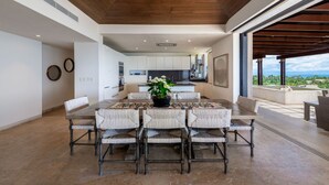 A modern kitchen with white cabinets and a central island, a dining area with a large table and woven chairs, and a view of the outdoors. - Vista Ballena 4BD - Tau Punta Mita (Punta de Mita)
