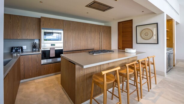 A modern kitchen with wooden cabinetry, a central island with bar stools, and built-in appliances. - Villa Cielo Azul 4BD - Iyari Punta Mita (Punta de Mita)