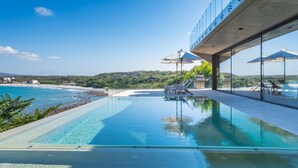 A modern outdoor swimming pool with clear blue water, surrounded by lounge chairs and umbrellas, overlooking a beach and hills. - Casa Morgandez 5BD - Iyari Punta Mita (Punta de Mita)