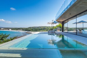 Ein moderner Außenpool mit klarem, blauem Wasser, umgeben von Liegestühlen und Sonnenschirmen, mit Blick auf einen Strand und Hügel.